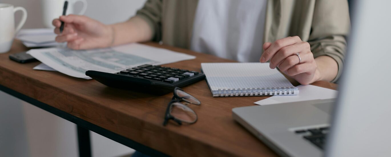A woman manages finances at home, using a laptop and calculator on a wooden desk.