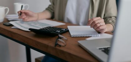 A woman manages finances at home, using a laptop and calculator on a wooden desk.