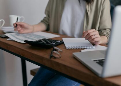 A woman manages finances at home, using a laptop and calculator on a wooden desk.